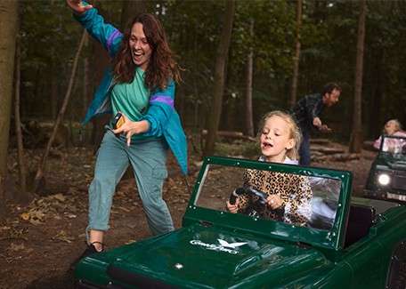 Child in a green toy jeep drives along a forest trail, smiling and gripping the wheel; an adult runs beside cheering, with other riders and trees in the background.