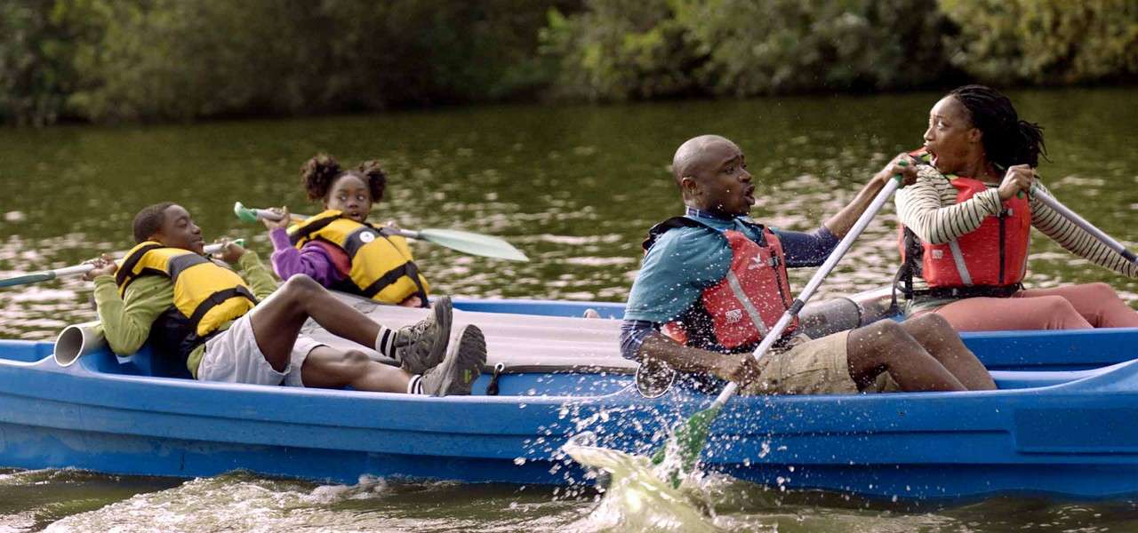 Four people in a blue rowboat paddle quickly, splashing water. Two adults row anxiously while two children recline with oars on a calm green lake bordered by trees.