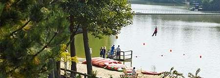 A person zip-lines over a lake, approaching a dock where people stand near kayaks; trees frame the sandy shore on a calm, sunny day.