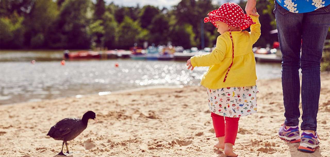 Child in yellow jacket and red polka-dot hat toddles barefoot, reaching toward a black waterbird; adult holds their hand. Context: sandy lakeshore with buoys, moored boats, and trees under daylight.