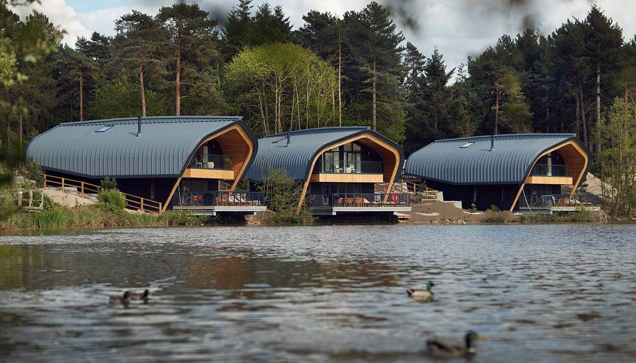 Three modern, curved-roof lodges overlook a lake, their glass fronts and decks raised on supports. Ducks glide across the foreground water, while dense conifer forest surrounds the shoreline.
