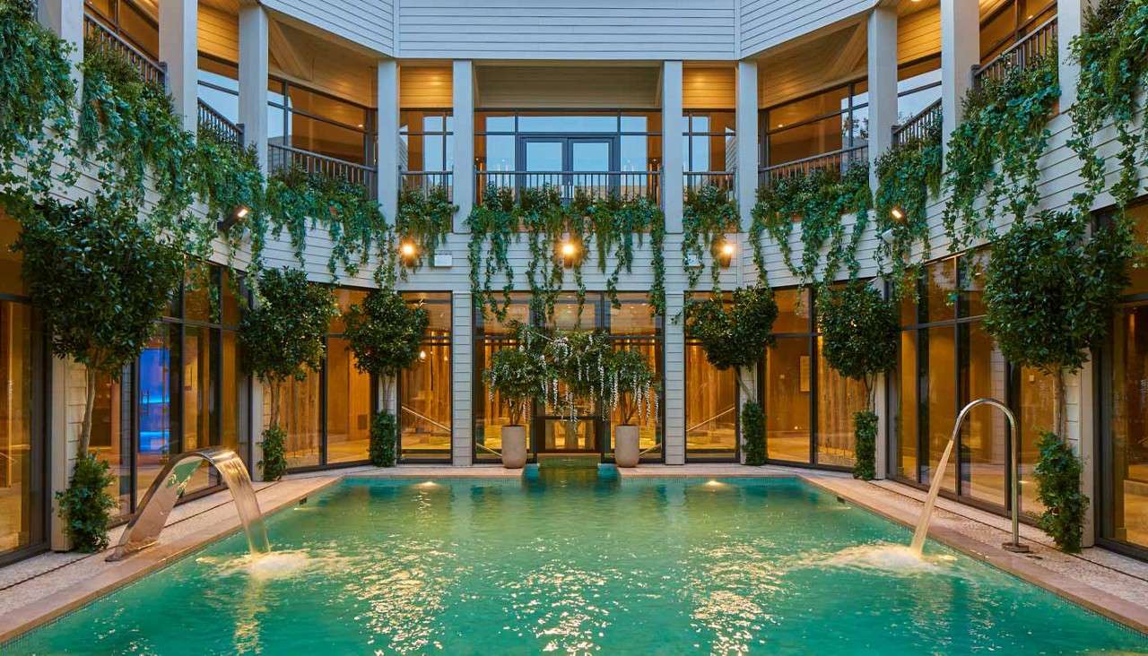 A turquoise swimming pool bubbles as two stainless spouts pour arcs of water. Surrounded by a two-story colonnade with glass walls, balconies, warm lights, and hanging vines and potted trees.