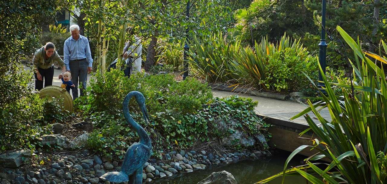 Family guides a toddler toward a small pond; the child reaches while adults support them. Nearby, a heron statue stands by rocks beside a wooden footbridge in a lush garden.