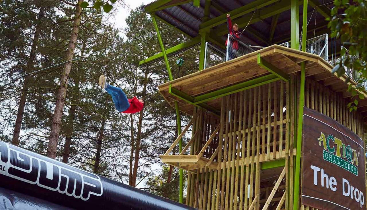 Person swings off a high wooden platform toward a large inflatable airbag in a forest adventure park. Visible text: JUMP; ACTION CHALLENGE; The Drop.