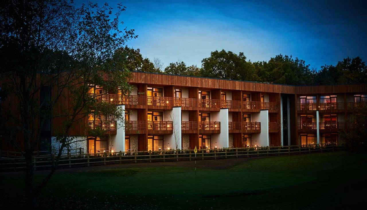 Hotel building glows with warm balcony lights, facing a manicured green with a flag. Evening sky darkens as trees surround the three-story, wood-clad structure behind a low fence.