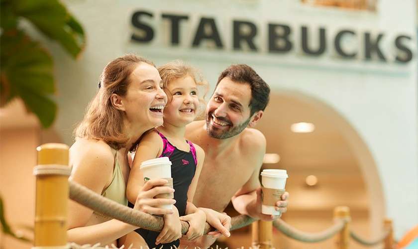 Two adults and a child smile while holding takeaway cups, leaning on a rope railing; indoor waterpark setting; background sign reads: STARBUCKS.