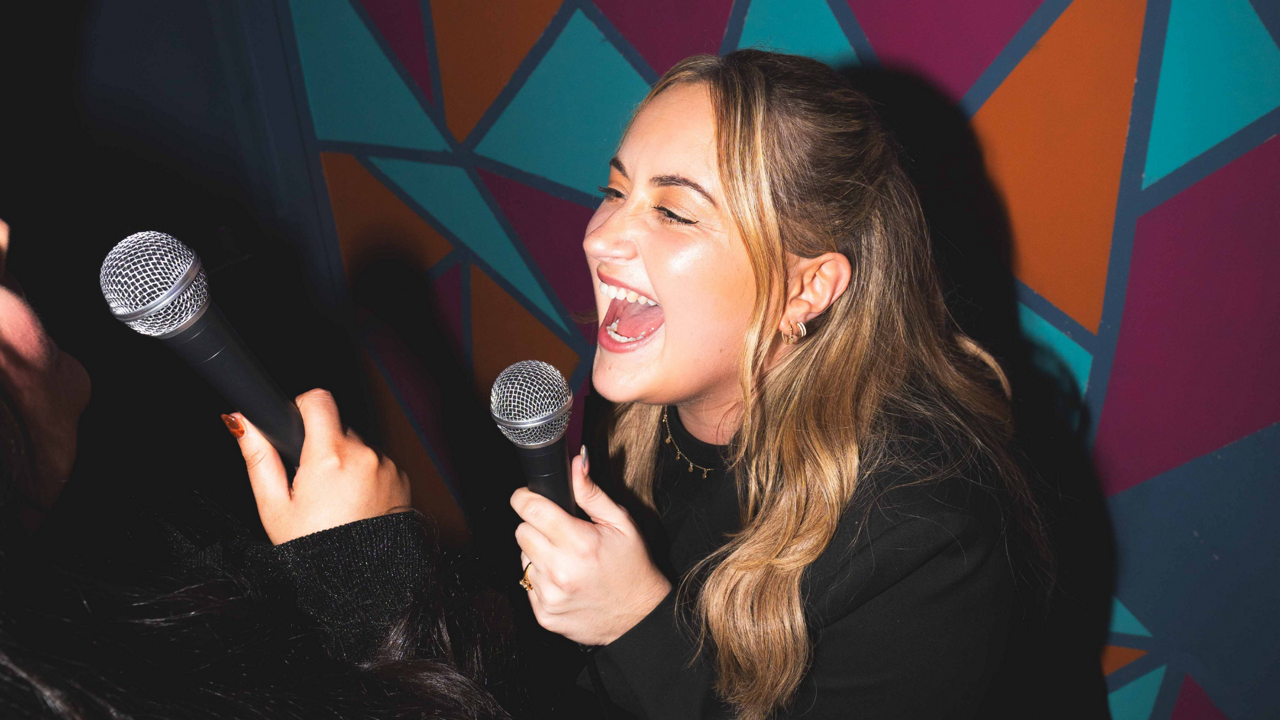 Smiling woman sings into a handheld microphone, mouth wide open, while another person holds a microphone nearby; both appear mid-song in a dim karaoke room with colorful geometric wall pattern.