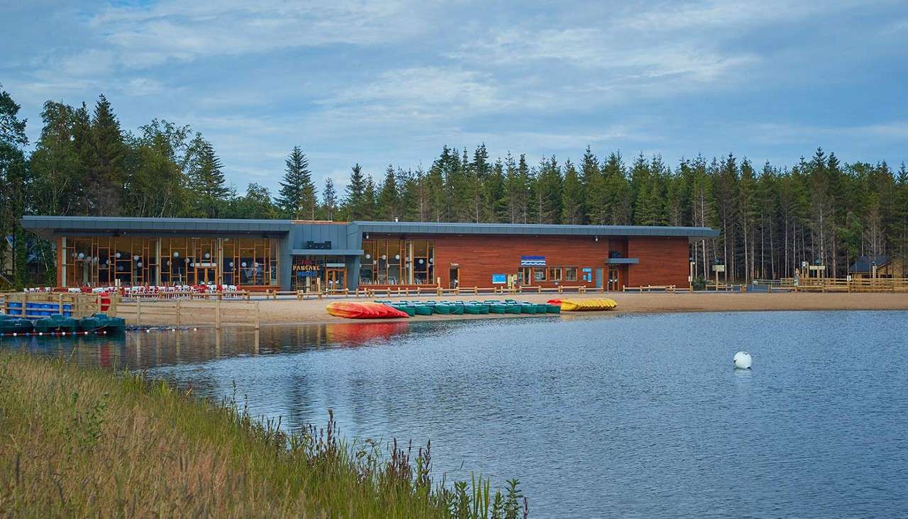 Watersports center building faces a sandy lakeshore, with stacked kayaks and canoes beside a floating dock. Calm water and a buoy foreground the scene, backed by forest under cloudy sky.
