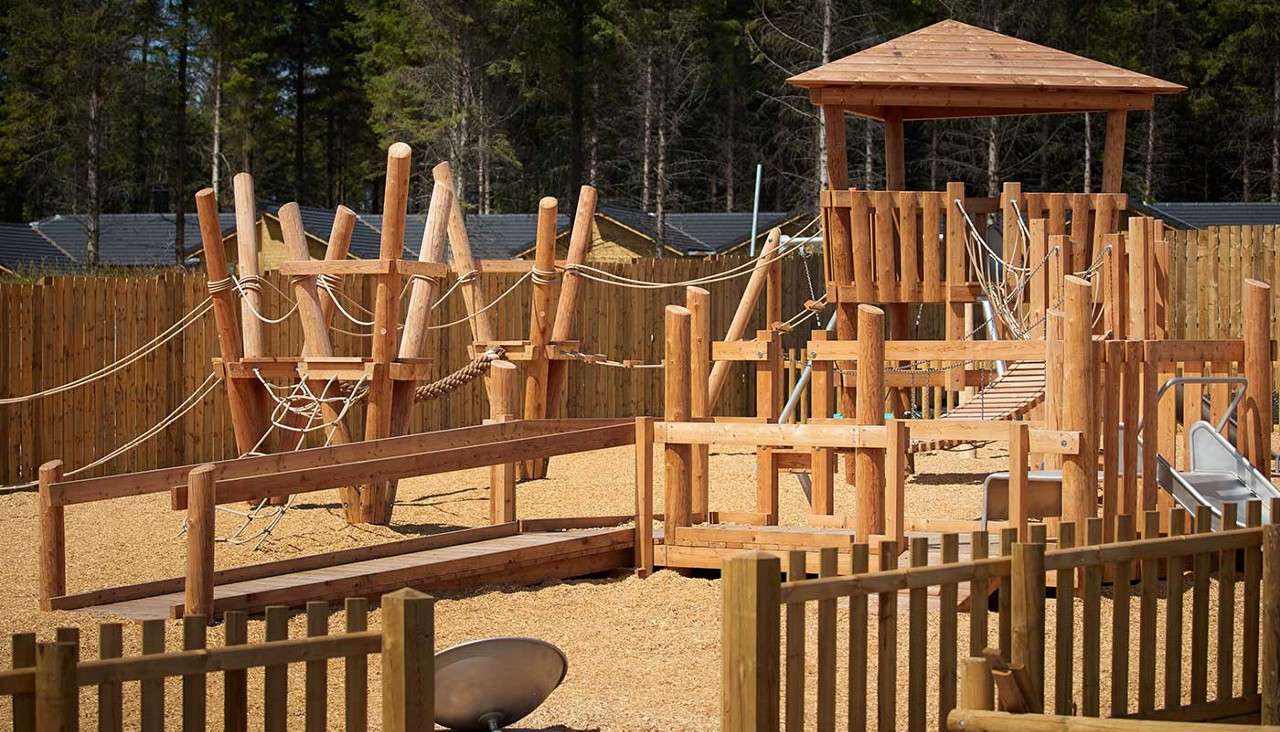 Wooden playground towers connect via rope bridges and ramps, enabling climbing and sliding; set on woodchip ground within a timber fence, with a small slide and forested backdrop.