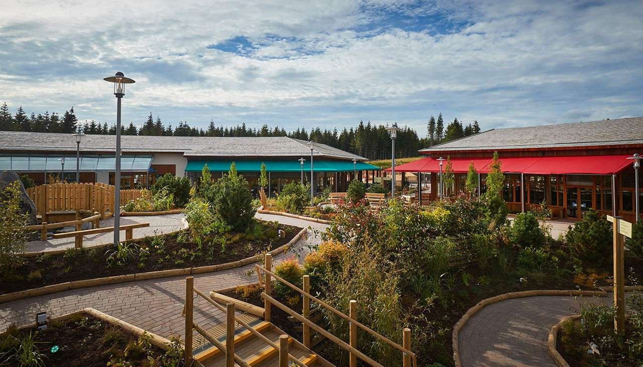 Garden paths wind through landscaped plant beds, guiding visitors between benches and lampposts, as single-story buildings with teal and red awnings surround the courtyard, set against a forested horizon under cloudy sky.