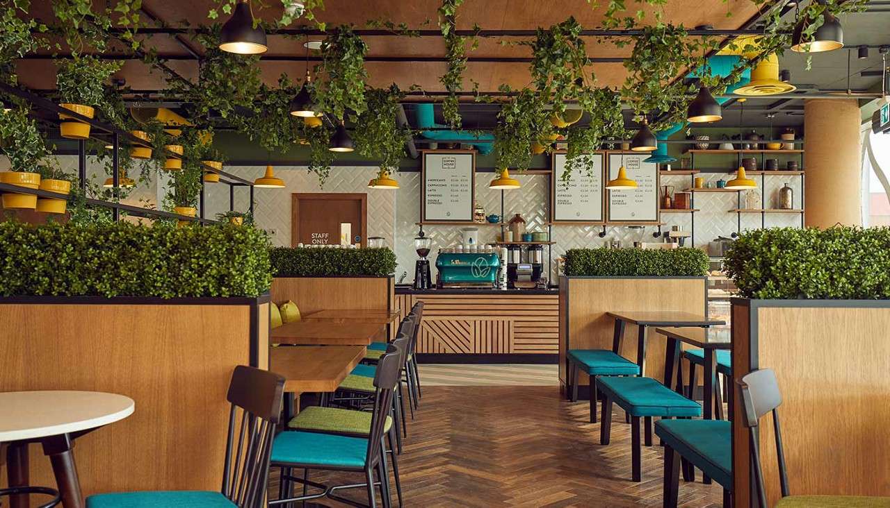 Cafe interior—plants hang from ceiling; pendant lamps light teal-seated booths; central counter operates teal espresso machine; wall shelves hold jars. Visible text: STAFF ONLY. Menu boards with drink lists and prices [illegible].