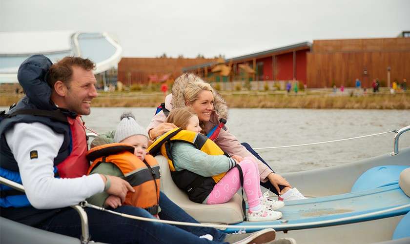 Family wearing life jackets rides a small pedal boat, smiling and seated together; in the background, modern wooden buildings and a waterside walkway line a lake under overcast sky.