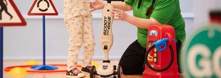 Child rides a three-wheeled scooter while an adult steadies it, in an indoor play area with toy traffic signs and cones; scooter text: "SCOOT AND RIDE"; green sign reads "GO".