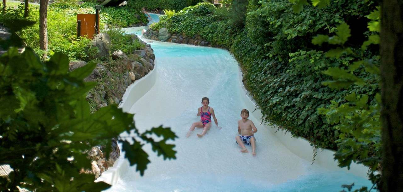 Two children slide feet-first down a curving shallow water slide, splashing lightly, surrounded by dense green foliage, trees, and rocks in an outdoor nature-themed water park.