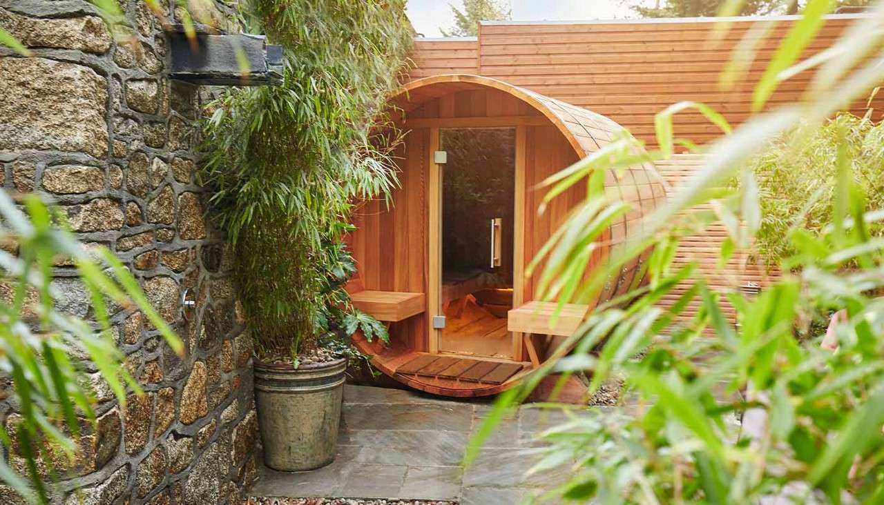 Wooden barrel-shaped sauna glows softly through a glass door, nestled between bamboo and a stone wall, with a slate path, potted plant, and small benches at the entrance.