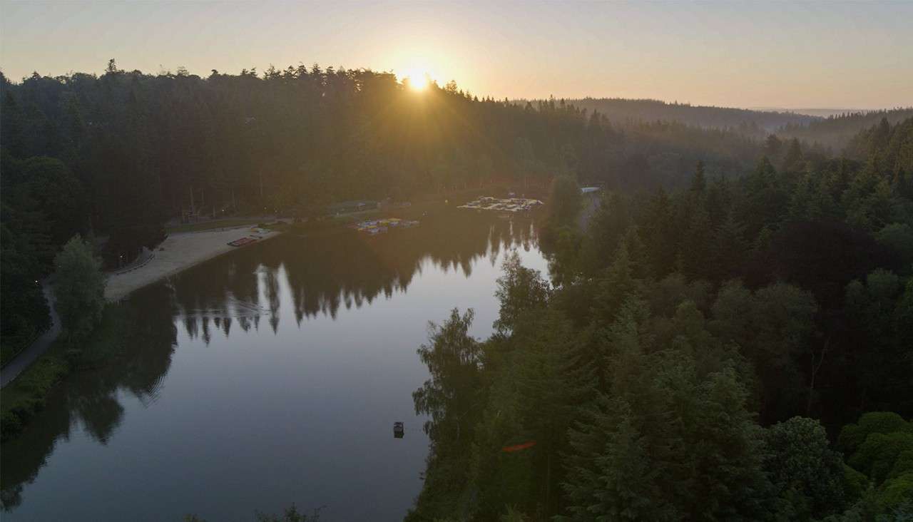 Lake reflects rising sun, bordered by dense evergreen forest; small beach and clustered boats sit along shore in soft morning haze, with distant hills fading into the horizon.