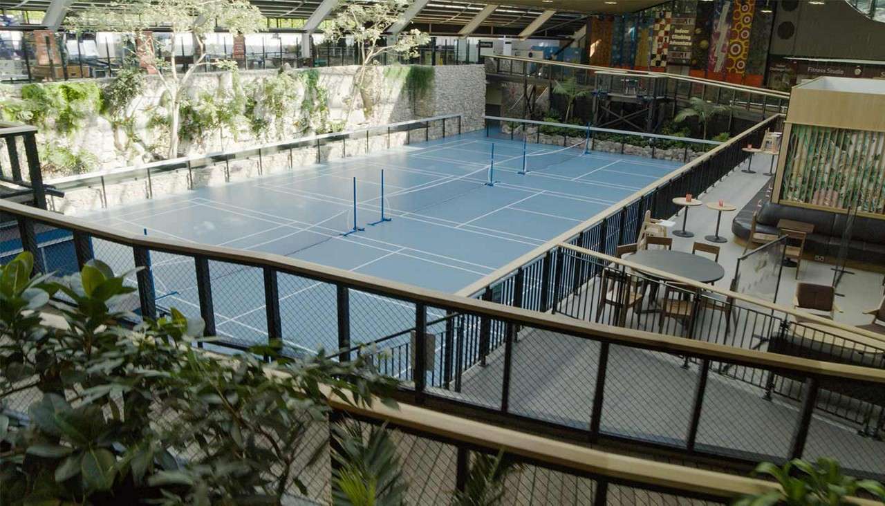 Empty blue pickleball courts sit unused within an indoor atrium; low nets stand upright. Surrounding balconies, plants, stone walls, skylights, and café tables create a leisure area overlooking the courts.