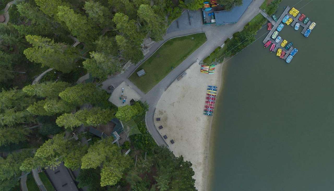 Colorful kayaks and paddle boats line a sandy lakeshore, resting beside calm water; forested park paths and docks surround the beach with gear storage and small buildings nearby.