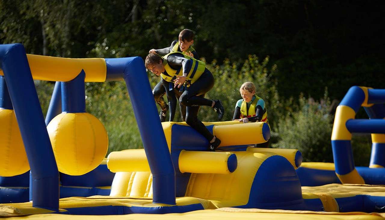 Three participants in wetsuits and yellow life vests scramble and jump over a blue-and-yellow inflatable obstacle course, amid large swinging balls, in an outdoor park with trees nearby.