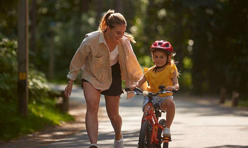 Child on a small red bicycle rides forward, wearing a helmet; adult jogs beside, steadying with a hand. Sunlit paved path curves through a wooded park.