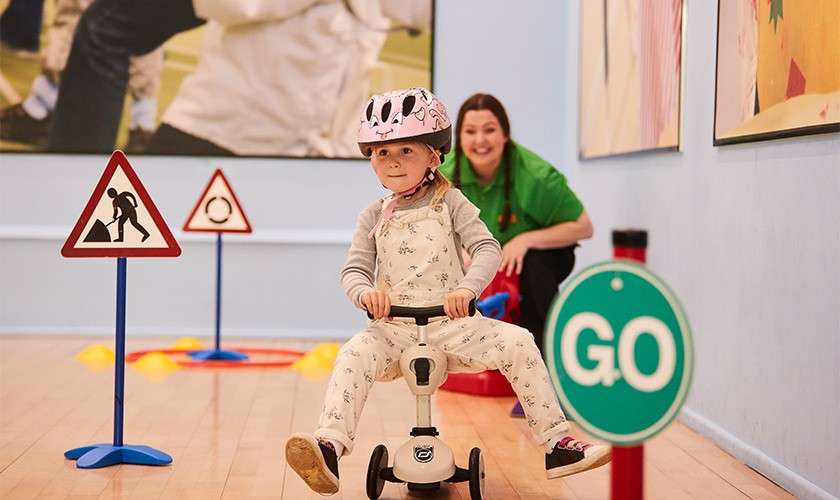 Child on a small tricycle rides forward, wearing a helmet, navigating an indoor play traffic course; an adult watches behind; cones and signs surround, including a green sign reading "GO".