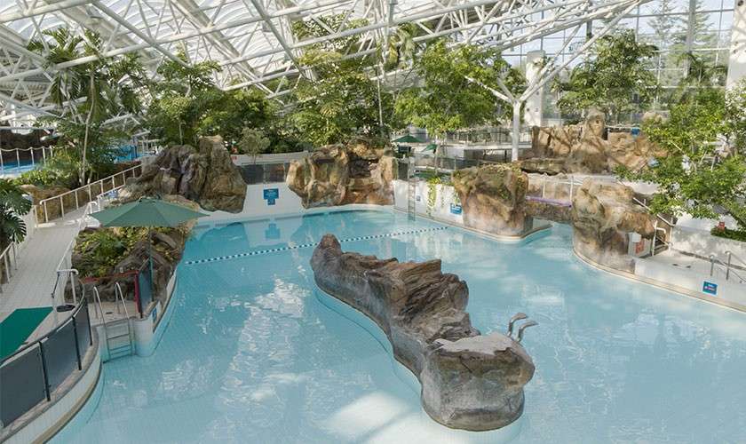 Indoor leisure pool curves around a central rock island, reflecting skylight; shallow, calm water surrounds artificial boulders, plants, and small slides within a glass-roofed atrium featuring walkways, railings, and umbrellas.