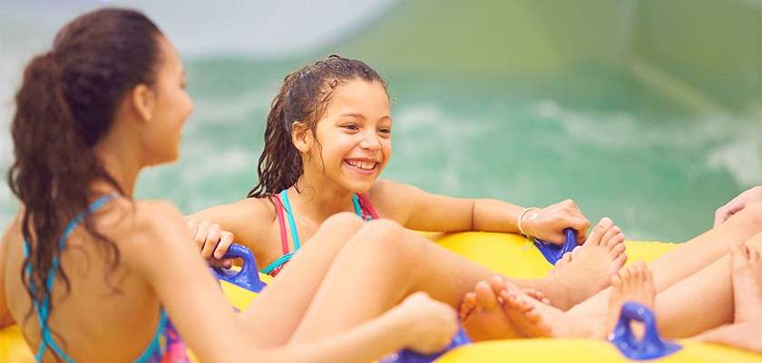 Children ride a yellow inflatable raft, laughing and holding handles, within an indoor water park, surrounded by greenish water and slide walls.