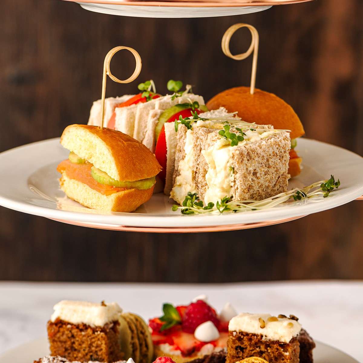 Assorted finger sandwiches sit neatly on a tiered stand, skewered and garnished with sprouts; below, blurred cakes and macarons suggest an afternoon tea spread against a dark wooden background.