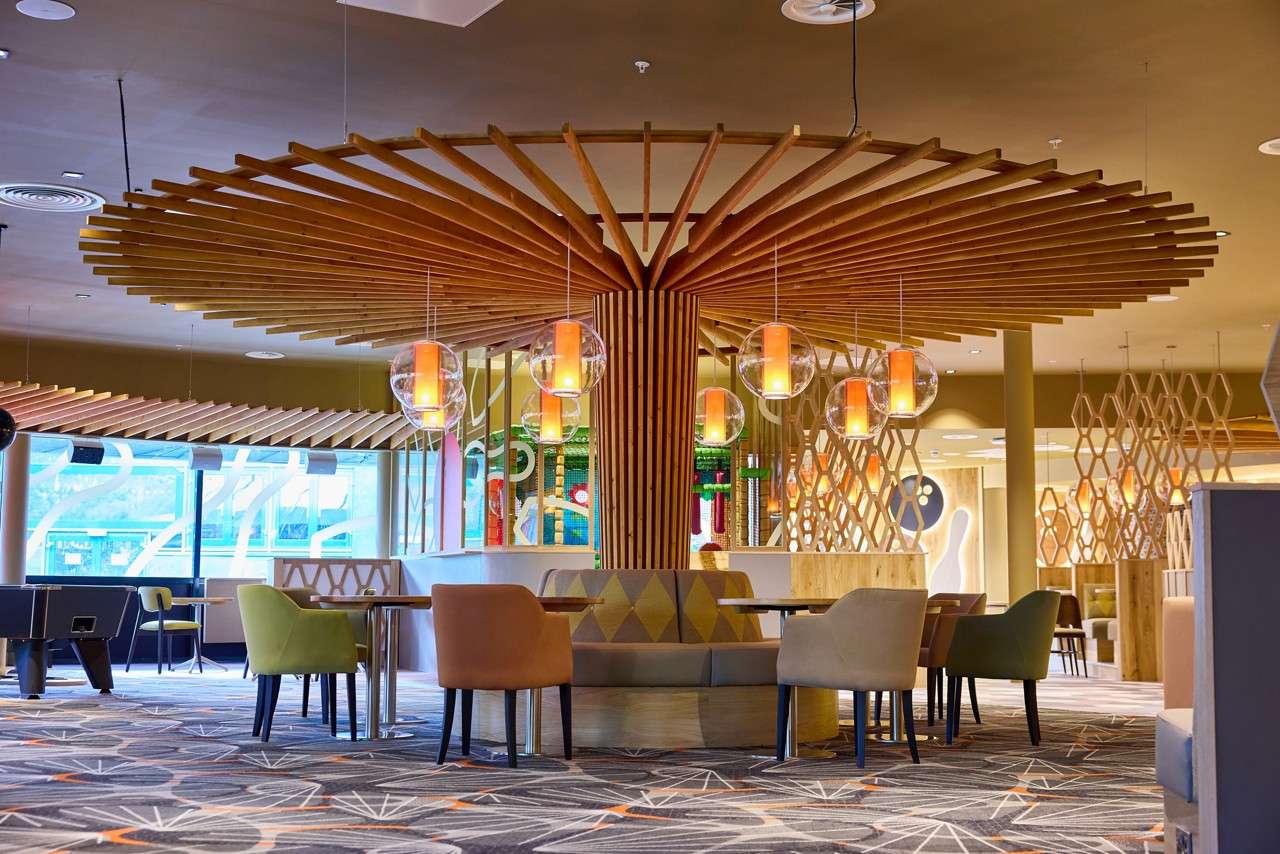 Circular booth seating surrounds a central wooden column; warm glass pendants hang above, illuminating scattered chairs and tables within a modern lounge featuring geometric partitions, patterned carpet, and large windows.