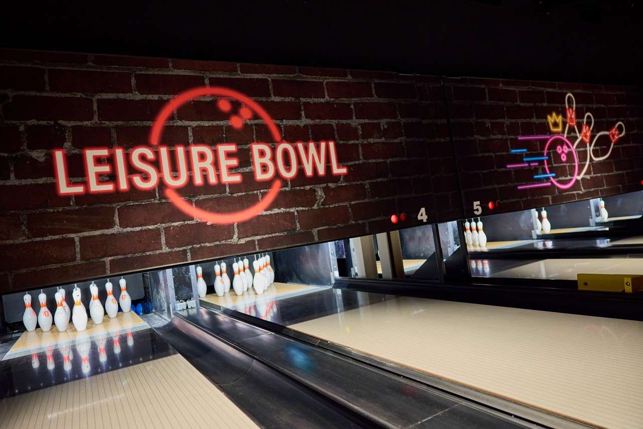 Bowling pins stand at lane ends, awaiting rolls, in a modern bowling alley; neon signage on brick reads “LEISURE BOWL”; lanes numbered 4 and 5 are visible.