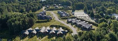 Wooden cabins cluster along a winding road, bordered by dense forest; additional buildings and rectangular courts sit nearby within a quiet, green hillside community with parking areas.