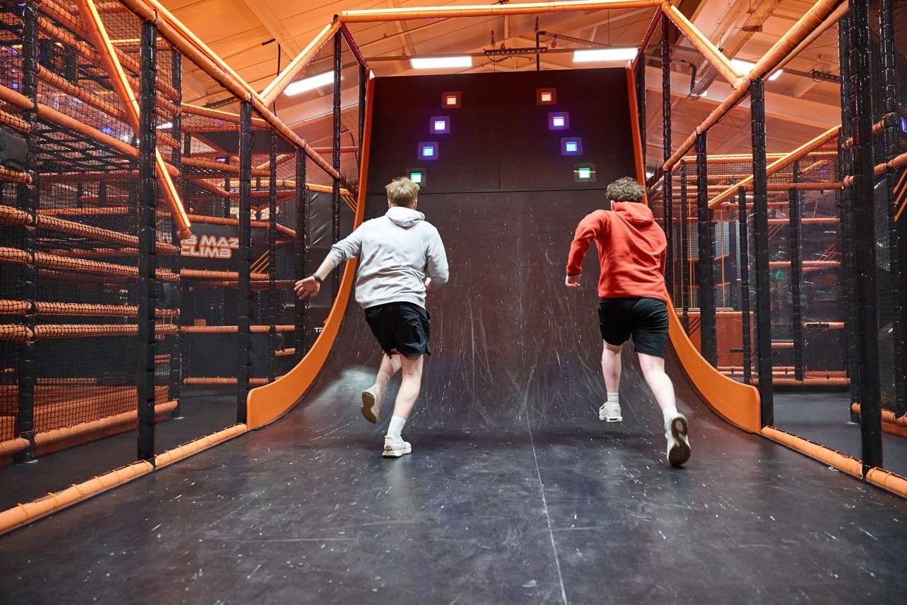 Two runners sprint up a curved ramp toward illuminated squares, inside an orange-framed indoor obstacle course with netted sides. Visible text: “MAZ CLIMB”.