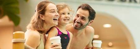 Three people smile and lean on a pool ledge; a child in a swimsuit between two adults holding takeaway cups, in a bright indoor aquatic area with plants and arched openings.