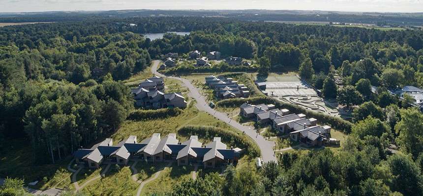 Cabin-style lodges line winding roads, arranged in clusters. They sit amid dense green forest, with parking areas, pathways, and a small lake in the distance under a bright sky.