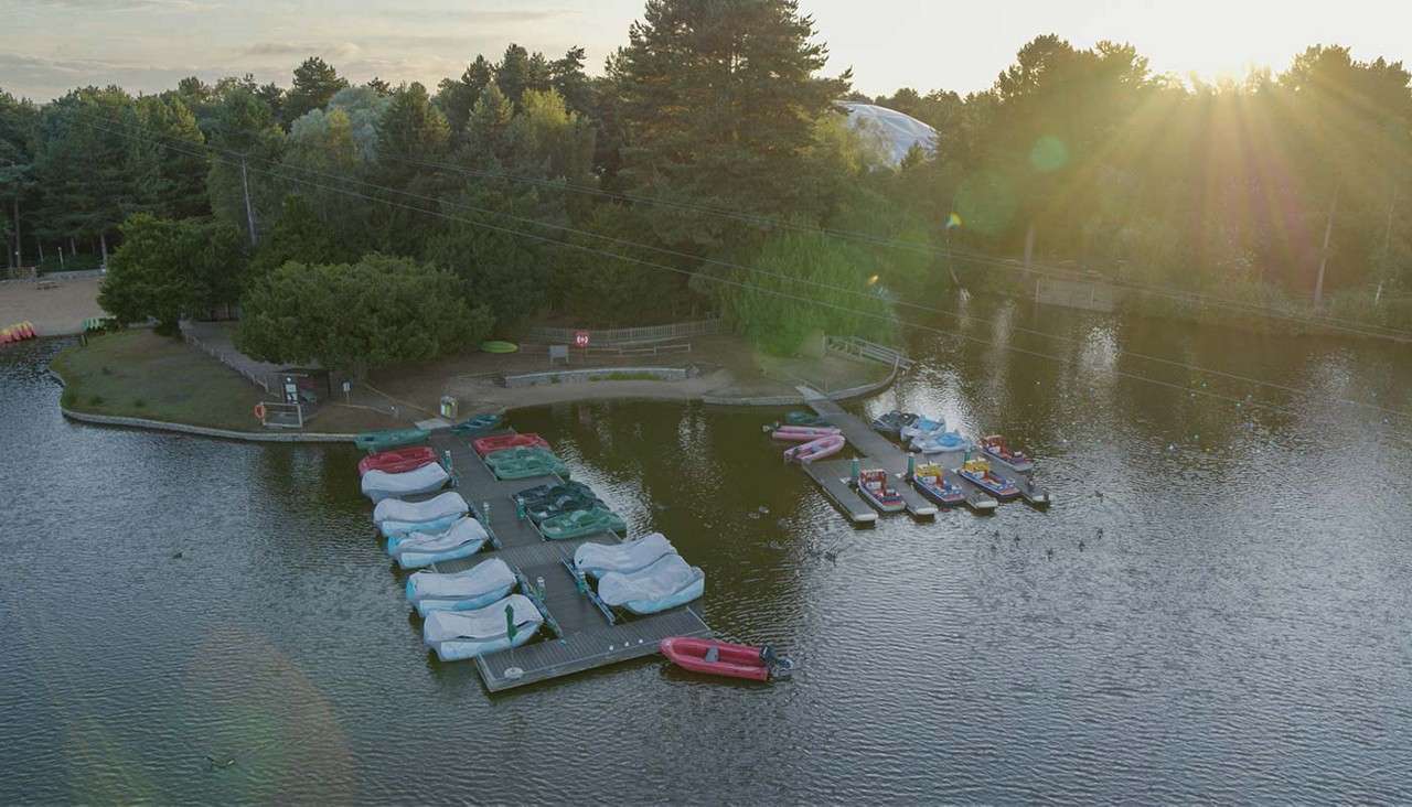 Paddle boats and small rental boats rest moored to wooden docks, while ducks drift nearby, in a calm forested lake at sunset, sunbeams and overhead cables spanning the water.