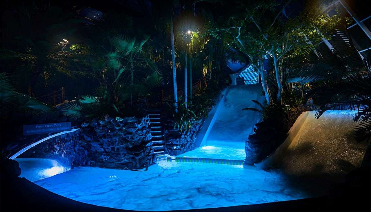 Waterslides discharge foaming water into a lit pool as runoff sheets down rock walls; tropical plants and stairs surround the scene in a dim, indoor setting. Text: “Feet First Only”.
