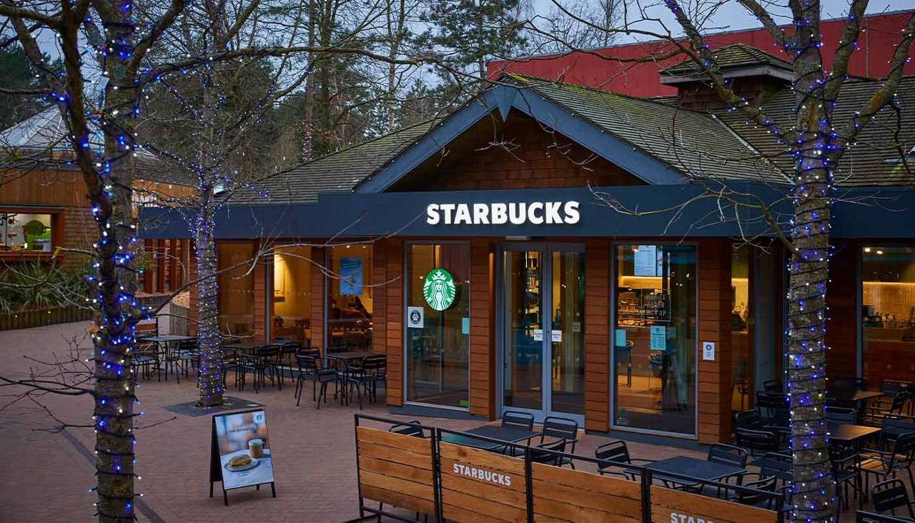 Starbucks café glows warmly, with customers inside and empty patio tables outside. Trees wrapped in blue lights frame the storefront in a pedestrian plaza. Text: STARBUCKS, STARBUCKS.