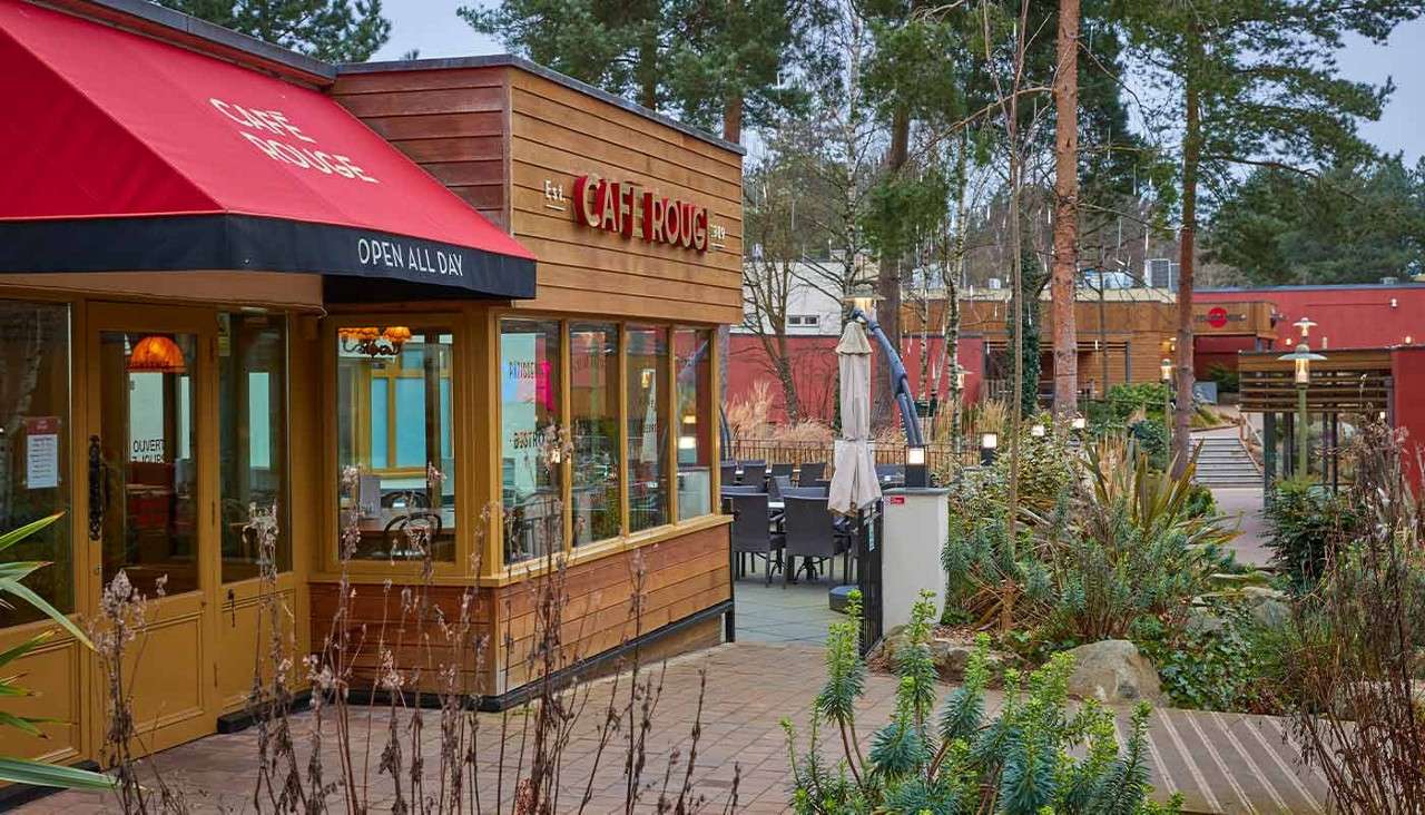Cafe exterior sits beside outdoor tables, within a landscaped pedestrian area with trees and plants. Text: OPEN ALL DAY; CAFE ROUGE; Est.; OUVERT; PATISSERIE; BISTRO.
