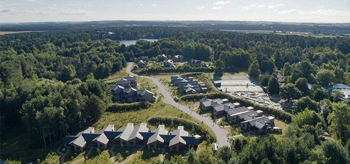 Clustered cabins sit amid dense forest, lining winding roads; nearby parking and pathways spread through the wooded landscape, with a distant lake and rolling countryside under bright sky.