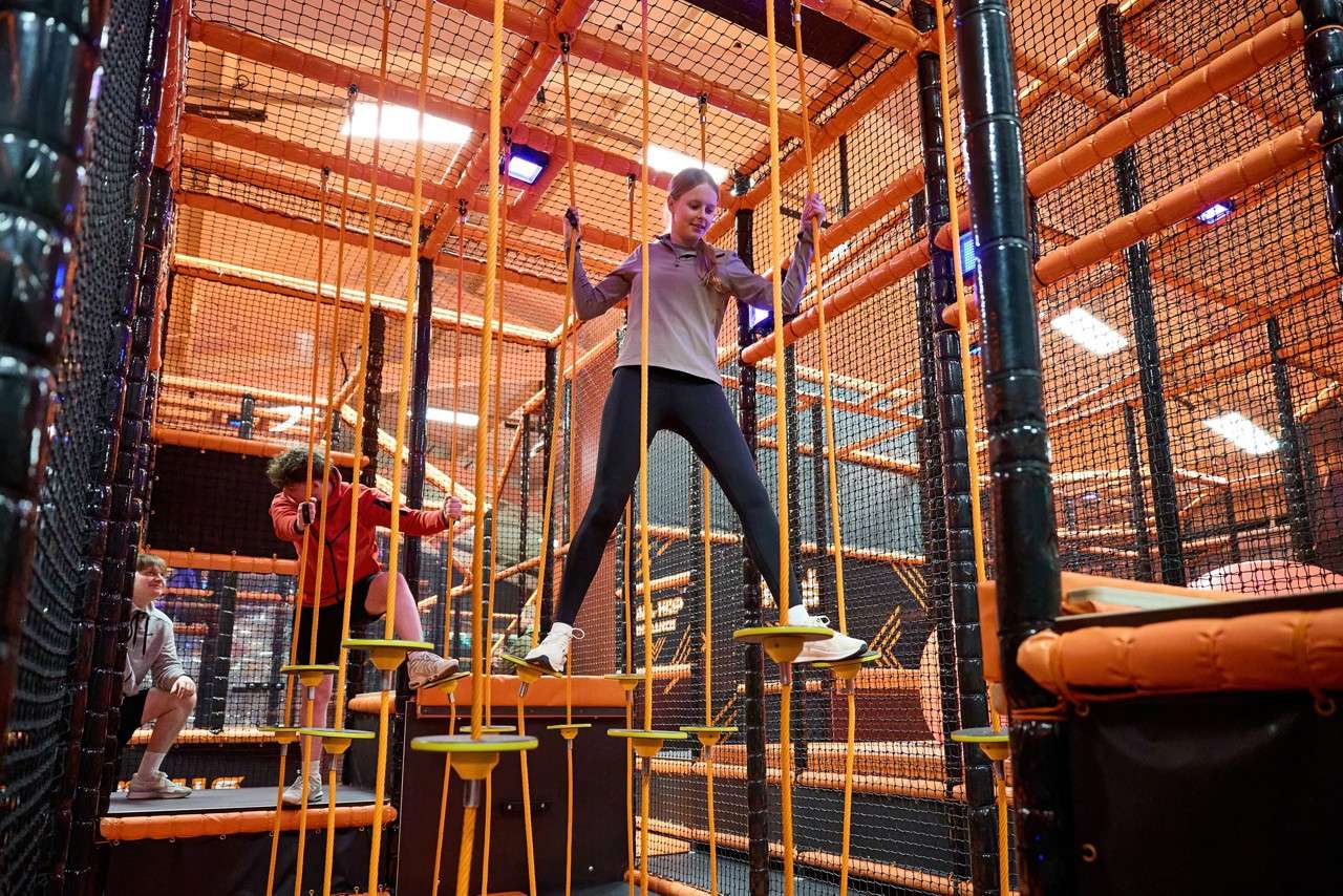Child balances on small suspended discs, gripping vertical ropes. Context: an indoor adventure course with orange frames and safety nets; two other children navigate and watch nearby.