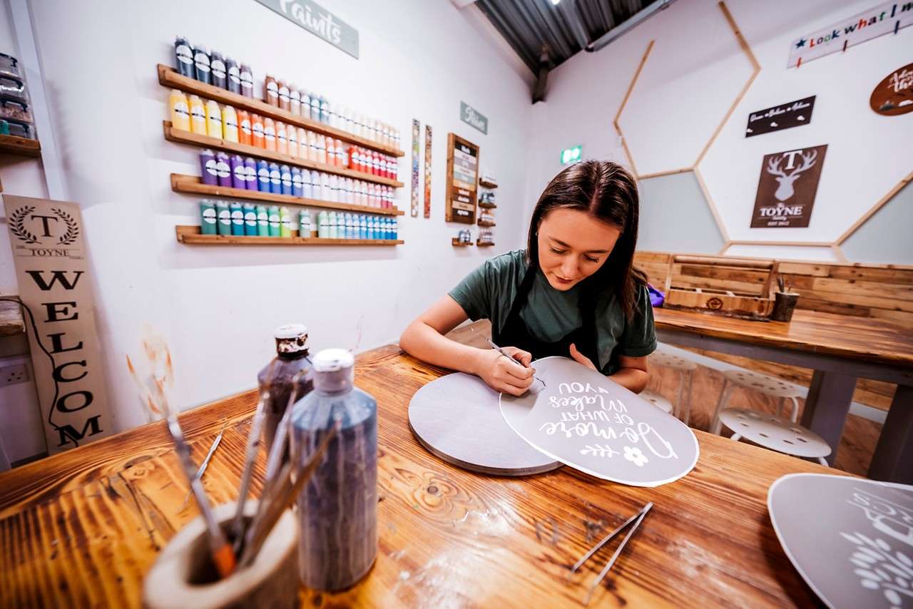 Woman paints lettering on a heart-shaped wooden sign in a craft studio. Text: “Be more of what makes you...”, “WELCOME”, “Paints”, “Stains”, “TOYNE”, “EXIT”.