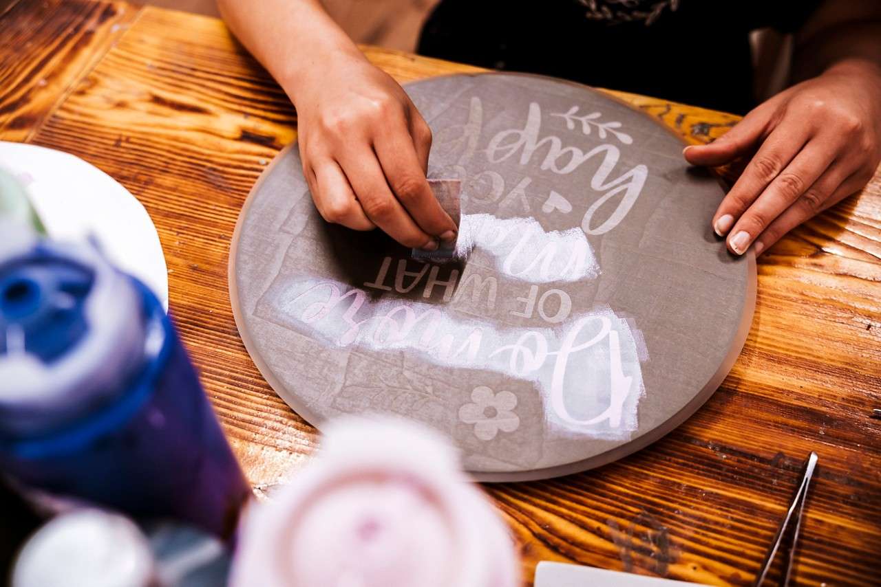 Hands rub a stencil onto a round gray board, transferring white letters with a squeegee on a wooden table with paint bottles. Text: "Do more of what makes you happy."