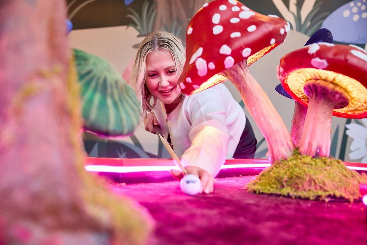 A person putts a white ball through oversized red-and-white mushrooms, on a mini-golf course surrounded by pink-purple turf and neon lighting in a whimsical indoor setting.