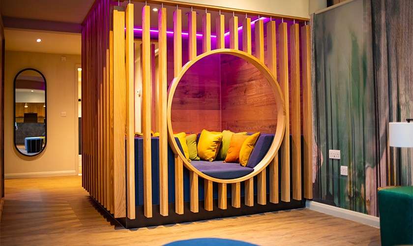 Circular wooden alcove seating cradles colorful cushions, illuminated by purple LED lighting, within a modern lounge; wood slats, blue bench, wood floors, wall mirror, abstract mural, and floor lamp surrounding.