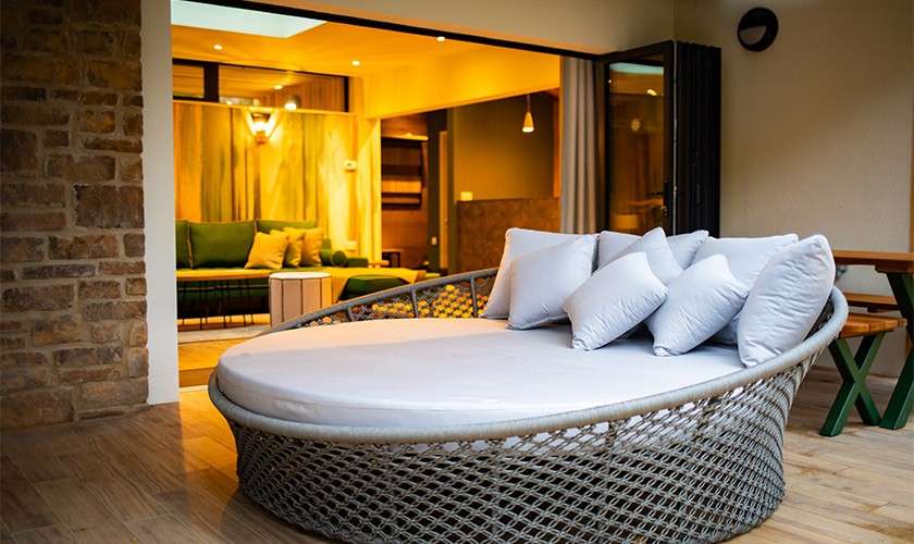 Round wicker daybed with white cushion sits on a wooden patio, framed by an open sliding doorway; inside, warm lighting reveals green sofas, a stone wall, and pendant lights.