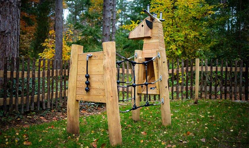 Wooden deer-shaped playground structure stands with rope-and-chain climbing net attached. Set on grass within a rustic fenced area, surrounded by tall trees and autumn foliage in a forested park.