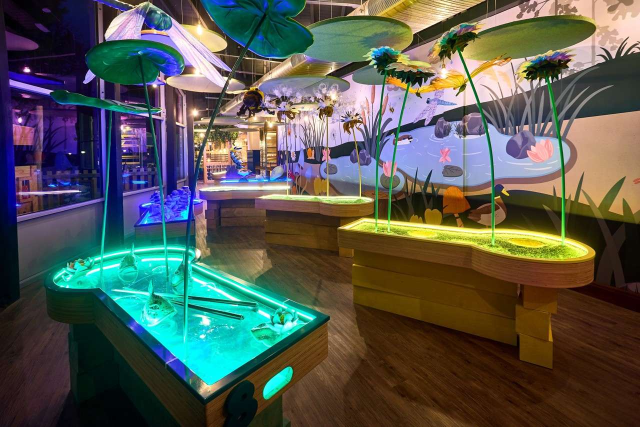 Glowing pond-shaped tables radiate turquoise and yellow, topped by oversized lily pads and flowers. Set in a dim indoor exhibit with a pond mural depicting ducks, rocks, and plants.