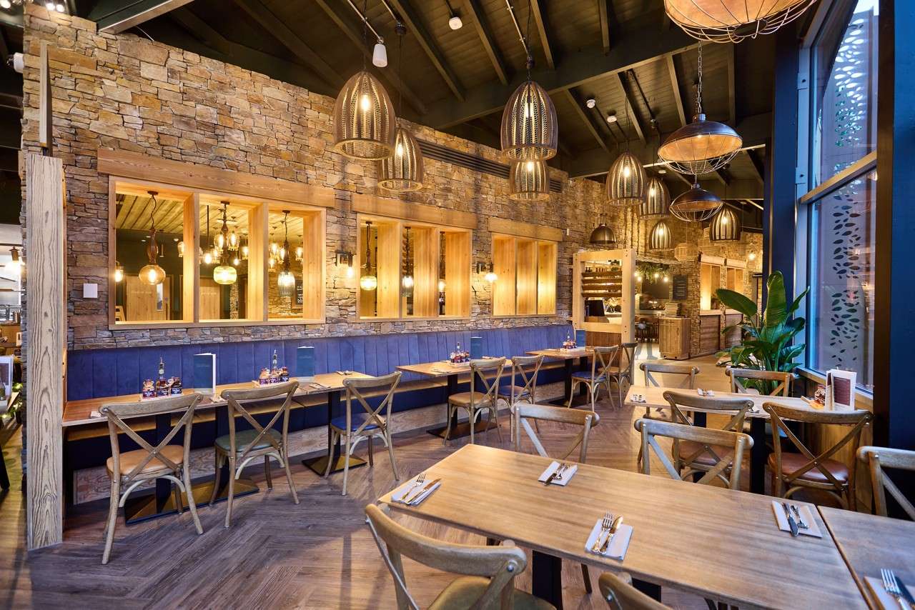 Tables and chairs sit neatly set with cutlery along a blue banquette. Pendant lights glow warmly, illuminating rustic stone walls, wooden accents, patterned floor, large windows, and a leafy plant.