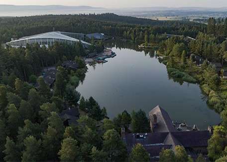 Lake reflects sky, bordered by dense pine forest; cabins and a glass-roofed complex line the shore. Aerial view shows paths and docks, with rolling hills and fields in the distance.