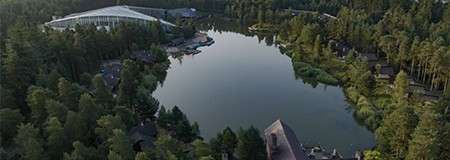 Lake reflects evening sky, bordered by dense forest. Aerial view shows scattered cabins, footpaths, and a large glass-roofed building near the shoreline, with calm water curving through a wooded resort.